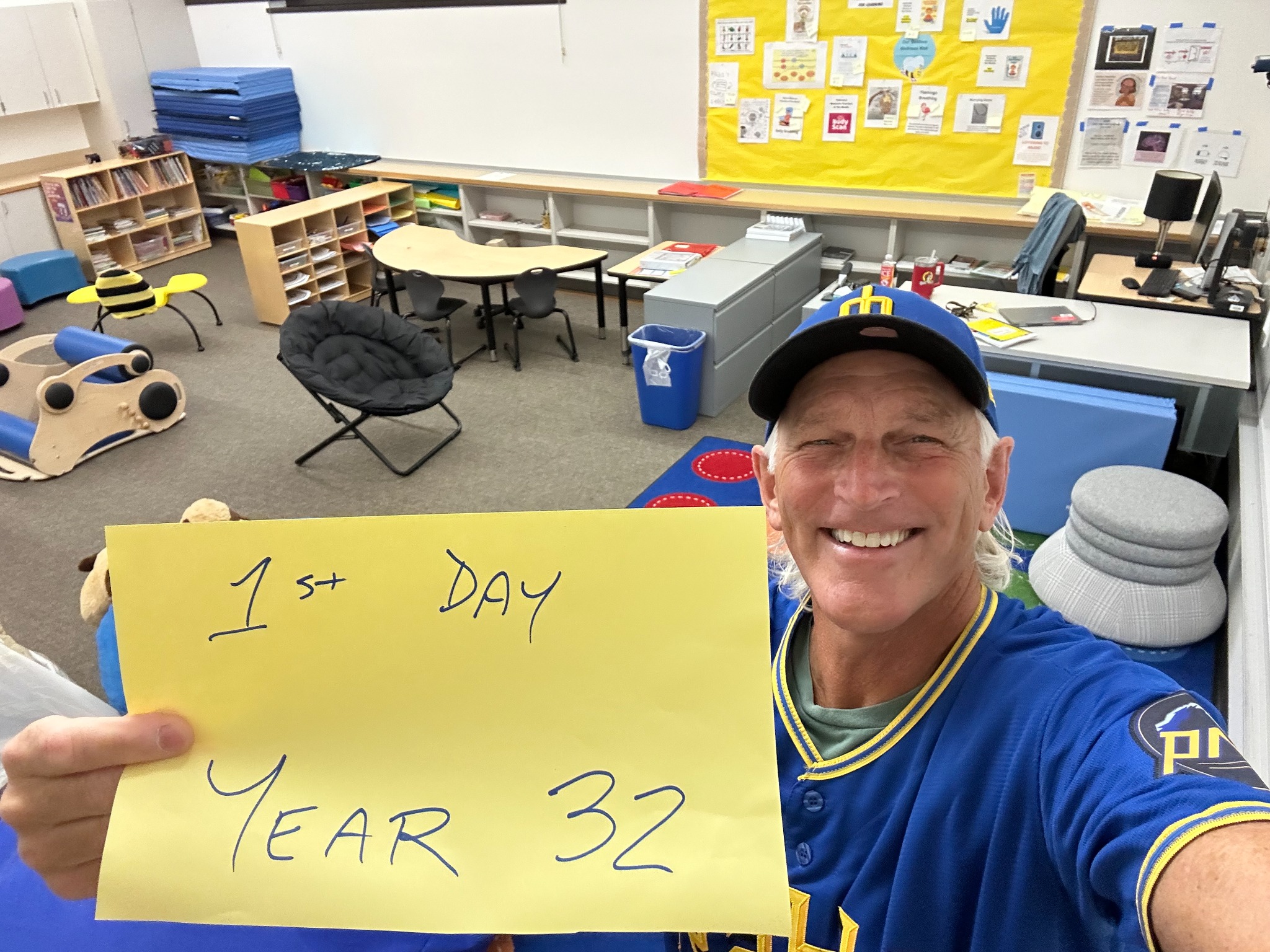 Jim Walters smiling in his classroom, holding a sign reading First Day, Year 32.